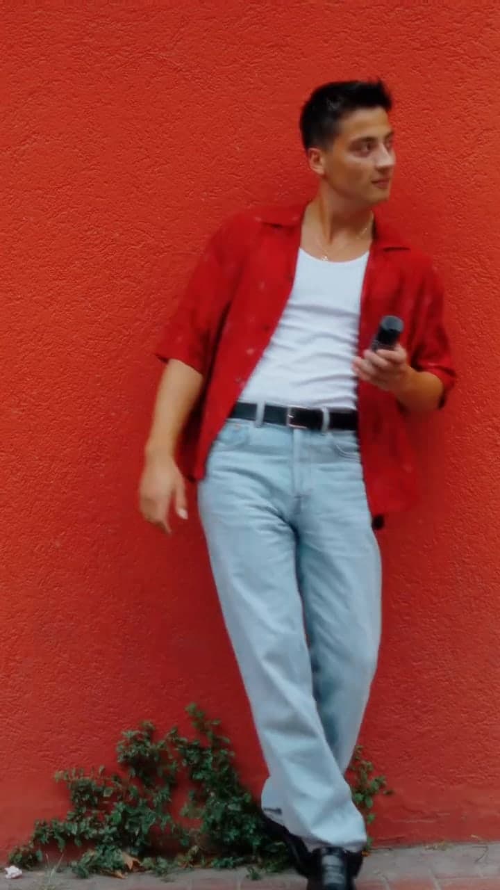 A man in an open red shirt and light jeans leaning against a red wall.
