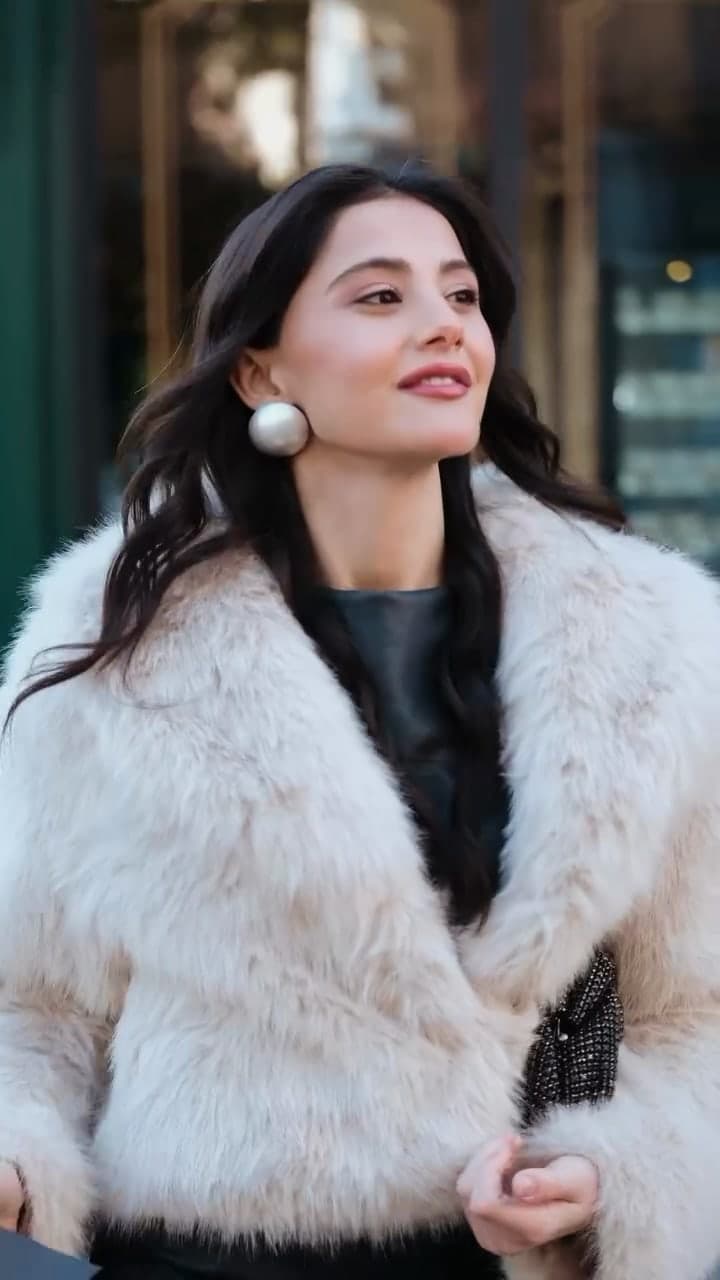 A woman in a cream fur jacket with pearl earrings on a city street.