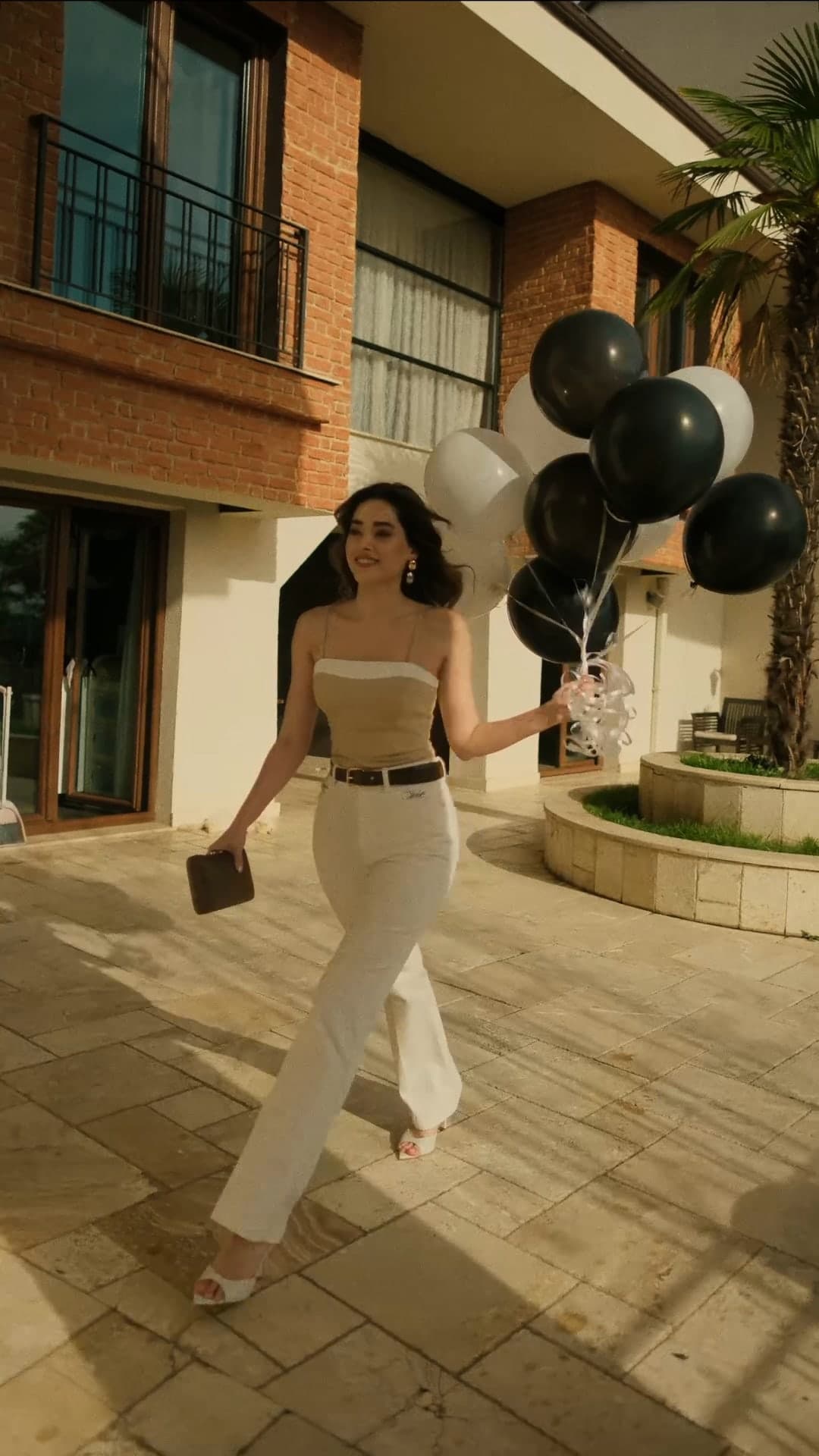 Simay Barlas in white trousers and a strapless top carrying black balloons.