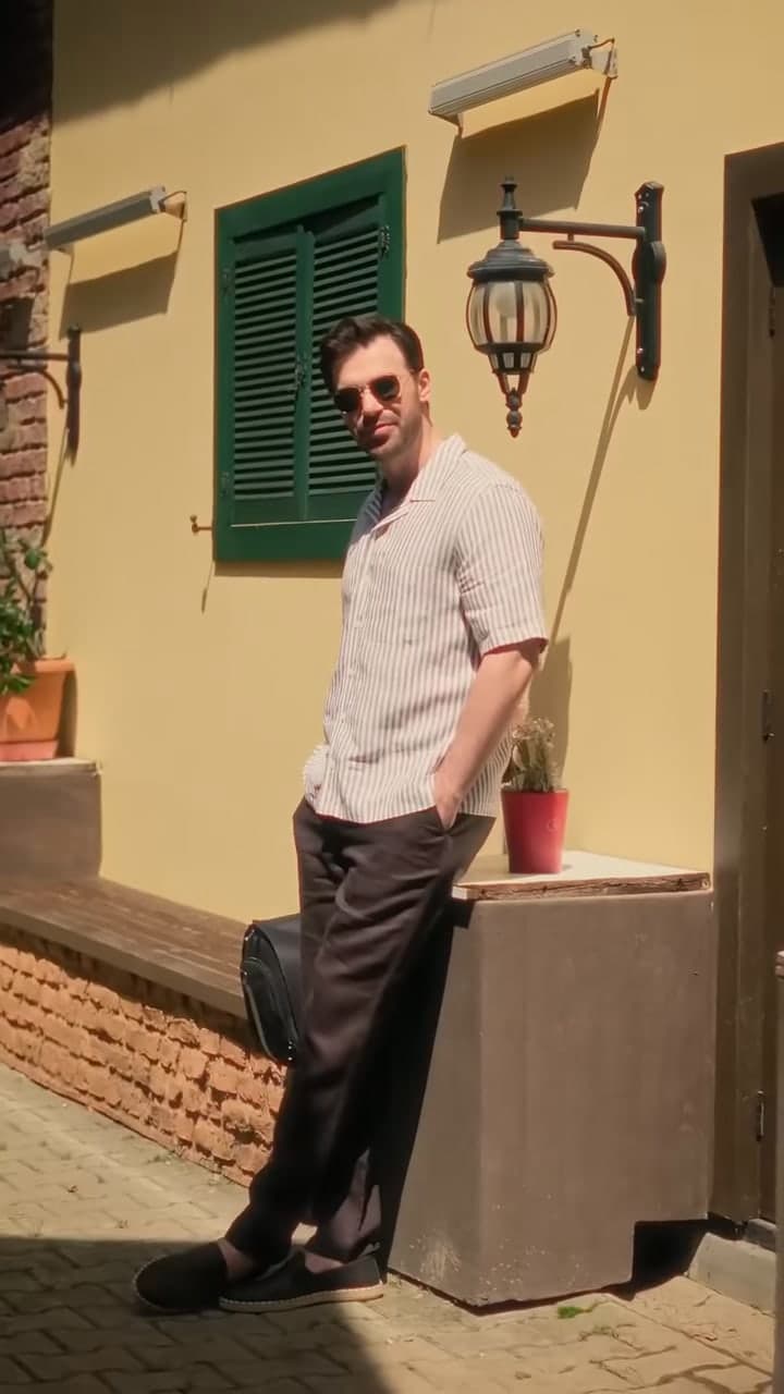 Yusuf Çim in a striped shirt leaning against a sunlit wall with green shutters.