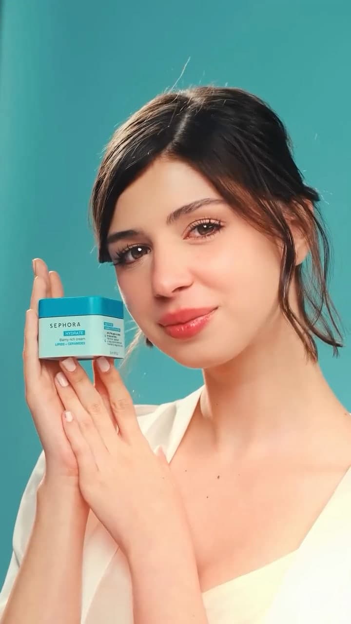 A woman in a white robe holding a Sephora Collection moisturiser against an aqua backdrop.