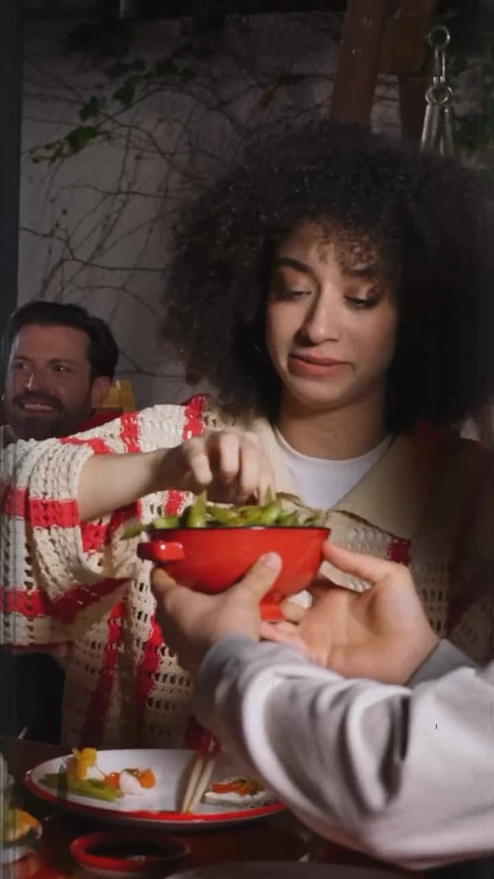 A woman with curly hair in a cream and red knit cardigan reaching for a bowl at a crowded table.