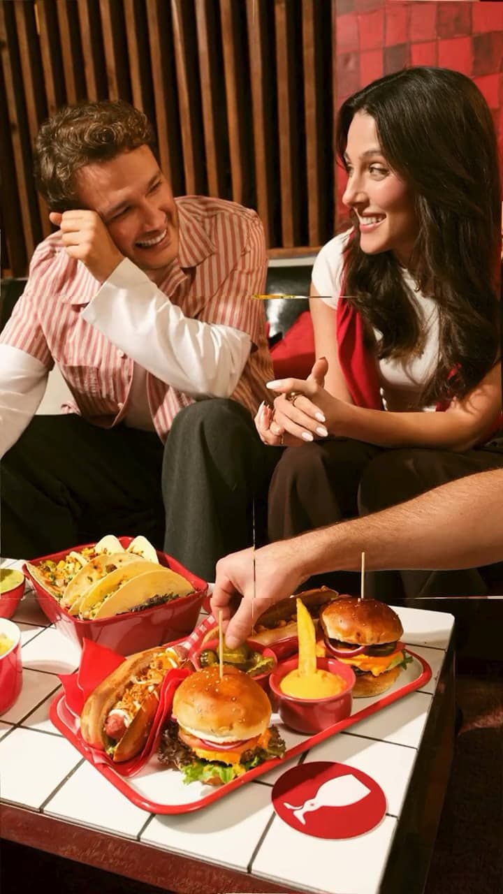 A man in a pinstriped shirt and a woman in a red vest sharing burgers and tacos.